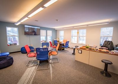 United Counseling Services in Bennington, Vermont. Photo description: A wide angle photo of PUCK's central space, well-lit by windows, showing tables with various therapeutic tools on them, colorful chairs, beanbags, bookcases full of books, a flat screen TV mounted on the wall, and other various items.