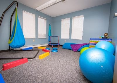 The sensory therapy room at PUCK. It is filled with a variety of therapeutic tools for helping children, especially those with sensory processing disorders. Photo description: A bright, colorful space filled with a swing, a trampoline, bouncy balls, a body stocking, and other toy-like items.