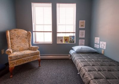 Quiet room at PUCK, United Counseling Services in Bennington Vermont. Photo description: The quiet room. There is an armchair to the left, and a comfortable small bed to the right. Two bright windows light up the room. Encouraging and motivational artwork decorates the wall on the space by the bed. There are books on the windowsill by the bed.