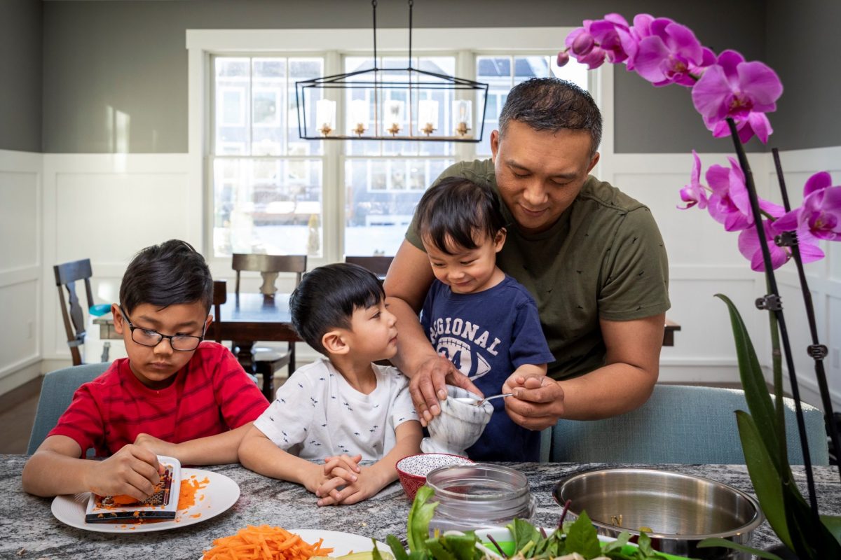 Pictured above / image description: A family prepares the ingredients for a healthy meal together. The young child on the left is shredding carrots, and a parent is supervising their two other children. In the forefront are salad greens and other ingredients in bowls and plates.