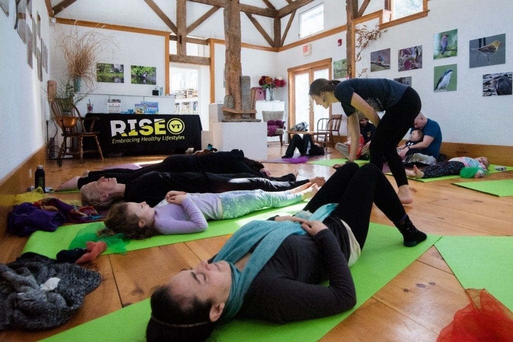 Children and parents lay on green yoga mats while the yoga teacher walks around giving suggestions for improving their poses. In the background there is a table with a RiseVT tablecloth in a large room. 