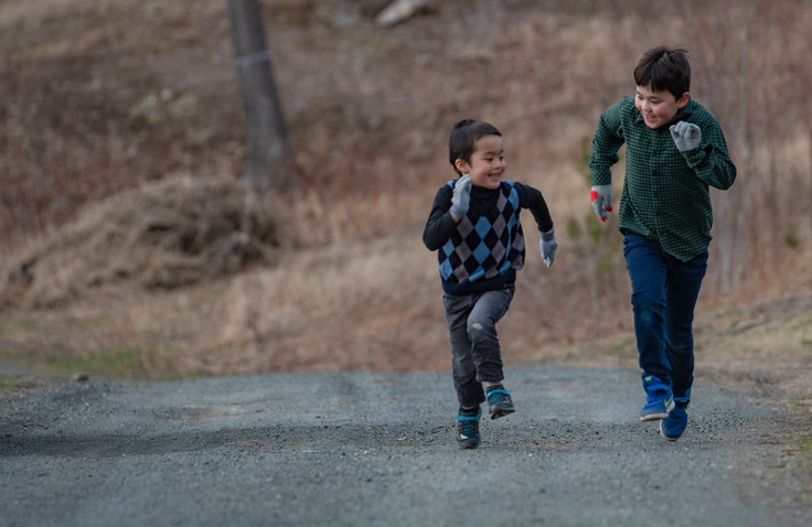 Two brothers are running up a dirt road together in the fall. The one is smaller than the other. They are both smiling as they race each other. 