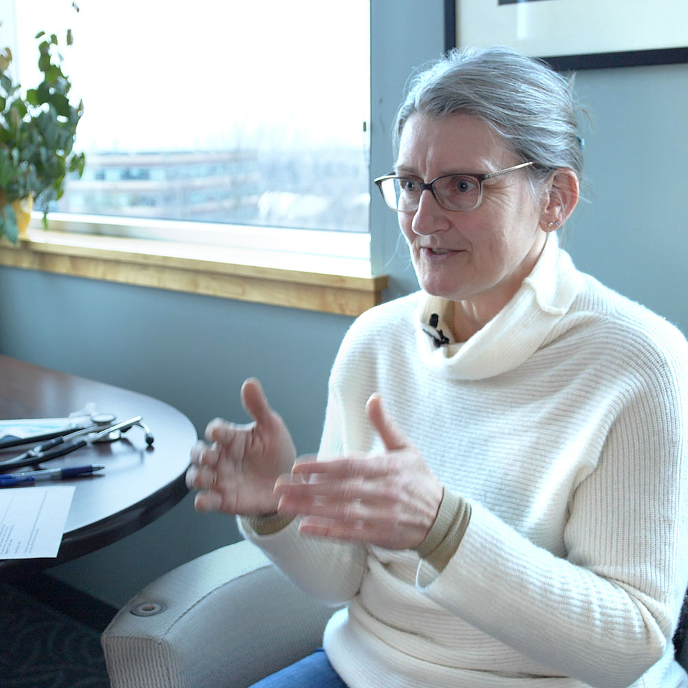 Photo of Dr. Alicia Jacobs, who has glasses and is wearing a white sweater. She is in an office and nearby a pair of stethoscopes are on a table.