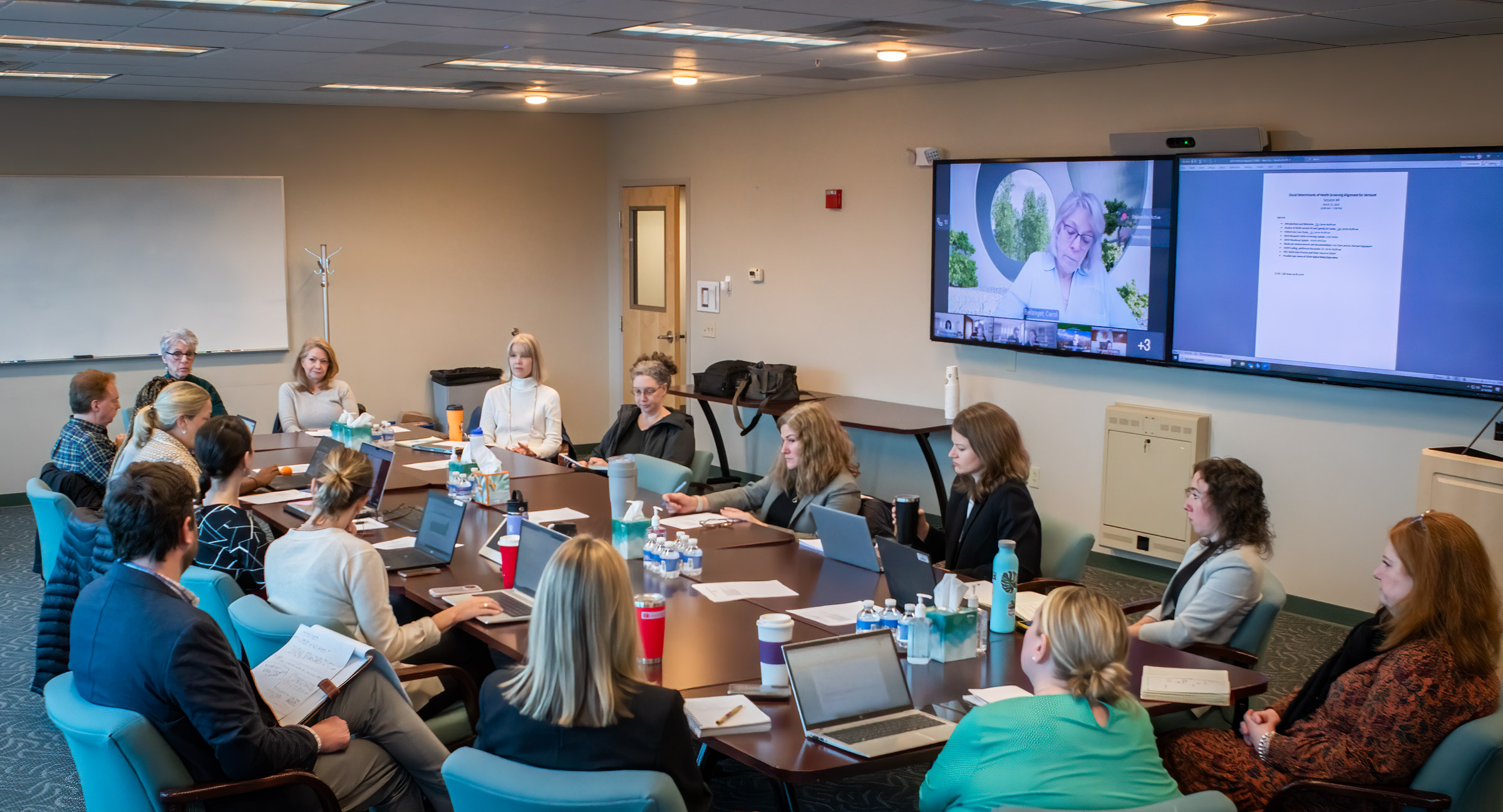 Members of the Social Determinants of Health meeting sit around a large conference room table. Behind them someone is calling in through the TV display via Microsoft Teams. 